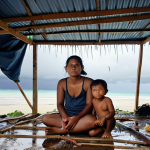 **Image Prompt:** A poignant photograph of Lani, a young Marshallese woman, sitting with her two children inside a makeshift shelter constructed from salvaged materials. Rain is leaking through the roof, highlighting the precariousness of their living situation. The background shows a blurred view of other similar shelters and signs of coastal erosion. Capture the vulnerability and resilience in their faces.